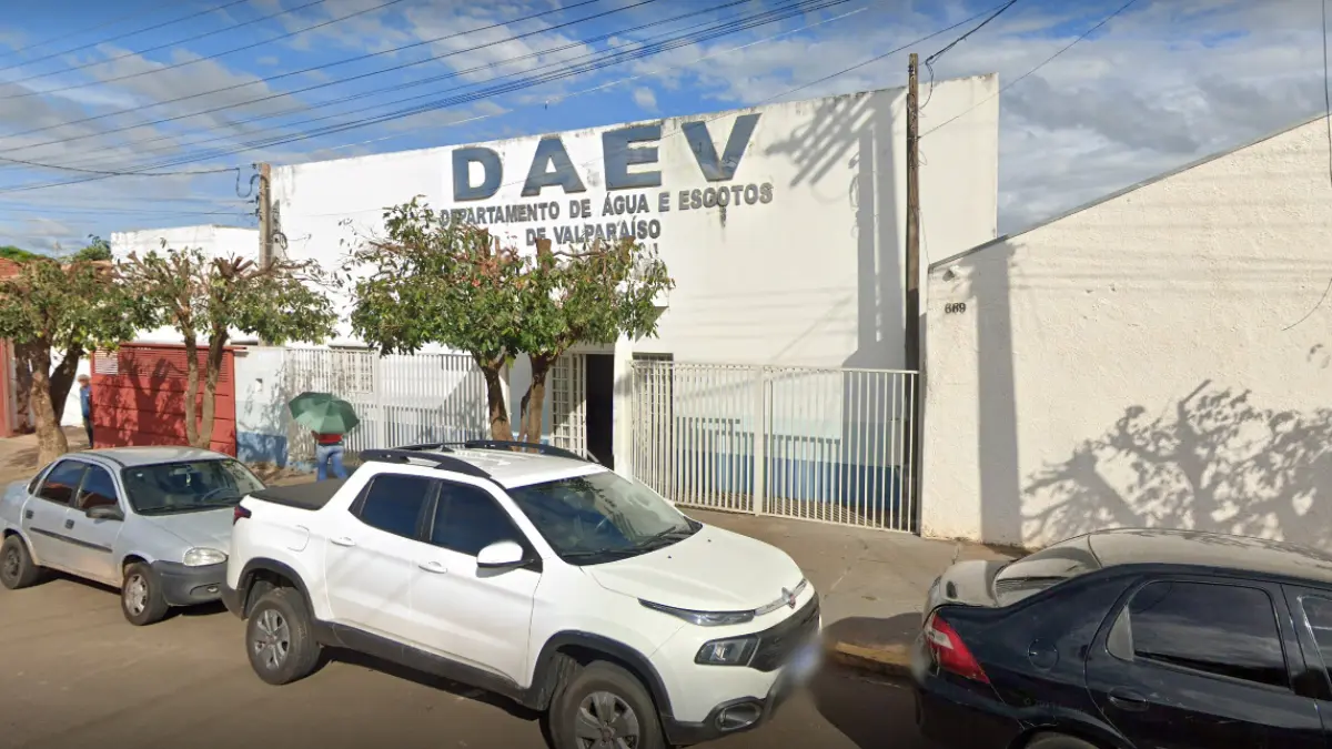 DAEV Valparaíso/SP divulga Resultado Preliminar do Concurso 2025 1 Fachada de um prédio branco com a inscrição "DAEV - Departamento de Água e Esgotos de Valparaíso". Há carros estacionados na frente e árvores de pequeno porte na calçada sob um céu com nuvens.