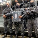 Agentes uniformizados do DEGASE-RJ posam em frente a viatura oficial com armamento e equipamento de proteção.
