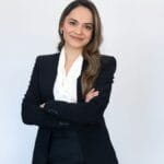 Mulher sorridente com cabelos castanhos longos, vestindo blazer preto e blusa branca, posa com os braços cruzados diante de fundo branco.