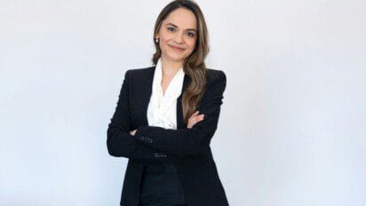 Mulher sorridente com cabelos castanhos longos, vestindo blazer preto e blusa branca, posa com os braços cruzados diante de fundo branco.