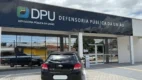 Fachada da Defensoria Pública da União (DPU) em Macapá, com letreiro preto e logotipo colorido, refletindo o céu e carros estacionados em frente ao prédio de arquitetura moderna e vidraças escuras.