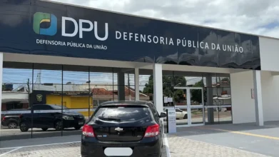 Fachada da Defensoria Pública da União (DPU) em Macapá, com letreiro preto e logotipo colorido, refletindo o céu e carros estacionados em frente ao prédio de arquitetura moderna e vidraças escuras.