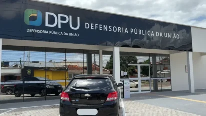 Fachada da Defensoria Pública da União (DPU) em Macapá, com letreiro preto e logotipo colorido, refletindo o céu e carros estacionados em frente ao prédio de arquitetura moderna e vidraças escuras.