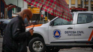 Processos Seletivos 6 Carro da Defesa Civil de Porto Alegre estacionado em dia de chuva com pessoa passando de guarda-chuva em primeiro plano.