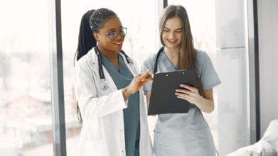 Processos Seletivos 7 Duas profissionais da saúde sorrindo enquanto analisam uma prancheta, vestindo jalecos e estetoscópios, em ambiente hospitalar iluminado.