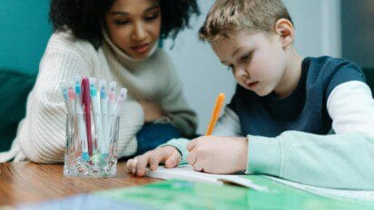 Mulher observando criança que escreve em caderno sobre mesa, com copo contendo canetas coloridas ao lado. Foto: cottonbro studio/Pexels