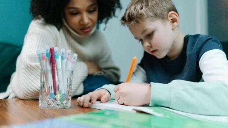 Mulher observando criança que escreve em caderno sobre mesa, com copo contendo canetas coloridas ao lado.