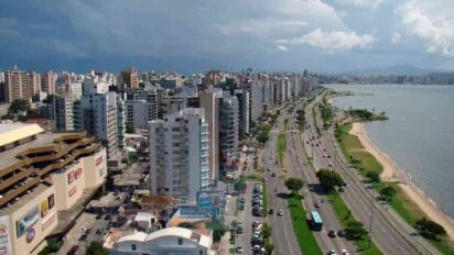 Vista aérea da orla de Florianópolis com prédios altos, avenida movimentada e faixa de praia ao lado do mar. Foto: Rodrigo Soldon/Wikipédia