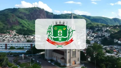 Vista aérea da cidade de Leopoldina, em Minas Gerais, com destaque para o brasão oficial do município centralizado sobre a imagem de uma catedral e montanhas ao fundo. Foto: Divulgação/Câmara Municipal de Leopoldina-MG (João Gabriel Baía Meneghite)