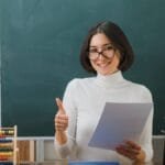 Professora sorridente segura papel e faz sinal de positivo em sala de aula com lousa ao fundo e materiais escolares sobre a mesa.