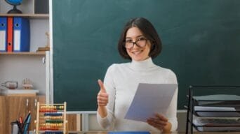Professora sorridente segura papel e faz sinal de positivo em sala de aula com lousa ao fundo e materiais escolares sobre a mesa.