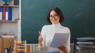 Processos Seletivos 5 Professora sorridente segura papel e faz sinal de positivo em sala de aula com lousa ao fundo e materiais escolares sobre a mesa.