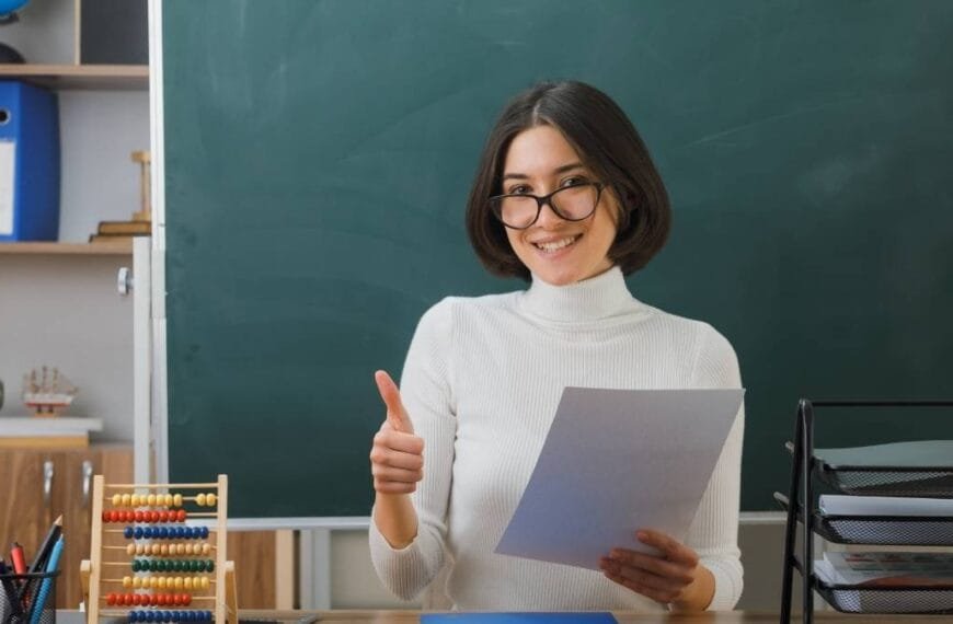 Professora sorridente segura papel e faz sinal de positivo em sala de aula com lousa ao fundo e materiais escolares sobre a mesa.