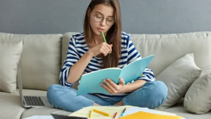 Mulher sentada em um sofá estudando, com caderno azul nas mãos, caneta verde na boca e laptop ao lado, concentrada na leitura e anotações. Foto: wayhomestudio/Freepik