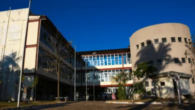 Fachada de prédio universitário com estrutura de vidro, paredes claras, mastros de bandeira e árvores sob céu azul, sede de especialização EaD gratuita