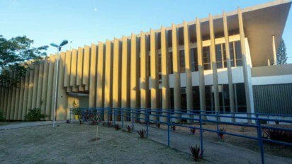 Fachada de um prédio moderno da UFAL com colunas verticais em destaque, rampa de acessibilidade azul, bicicletas estacionadas e céu claro ao fundo.