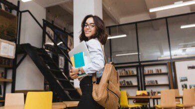 Jovem sorridente de óculos está em um ambiente moderno de biblioteca ou espaço de estudos (especialização). Ela carrega uma mochila bege nos ombros, segura um notebook prateado e cadernos coloridos nos braços. Usa camisa clara e calça escura, olhando para trás enquanto caminha. Ao fundo, há estantes com livros, mesas com cadeiras amarelas e uma escada que leva a um mezanino com mais quadros e iluminação artificial.