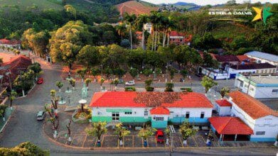 Vista aérea da cidade de Estrela Dalva-MG com destaque para a praça central arborizada, edifícios com telhados vermelhos e vegetação ao fundo.