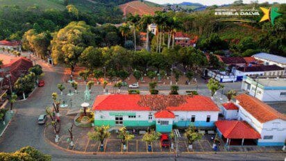 Vista aérea da cidade de Estrela Dalva-MG com destaque para a praça central arborizada, edifícios com telhados vermelhos e vegetação ao fundo.