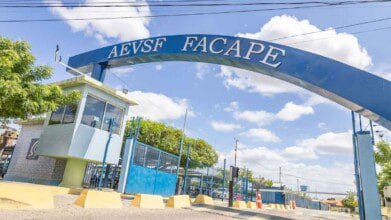 Processos Seletivos 3 Entrada principal da FACAPE em dia ensolarado, com arco azul metálico com o nome da instituição, guarita envidraçada, árvores, calçada e via de acesso com cones.