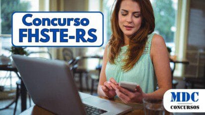 Mulher de cabelos ruivos e vestido verde-água sentada em um café, segurando um smartphone enquanto olha para a tela. Na mesa há um laptop aberto, uma xícara e um copo com água. O ambiente é iluminado, com grandes janelas ao fundo mostrando árvores e carros estacionados - FHSTE RS oferece 21 vagas e salários de até R$ 6.259 em Concurso