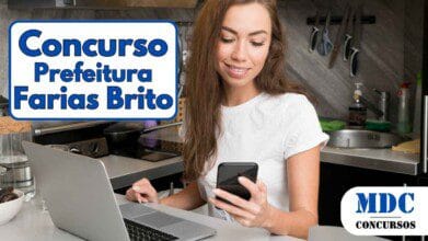 Mulher sorridente com cabelos longos e castanhos usa camiseta branca enquanto usa um notebook e segura um smartphone em uma cozinha moderna com armários amarelos - Farias Brito (CE) oferece 232 vagas e salários de até R$ 9,4 mil em Concurso