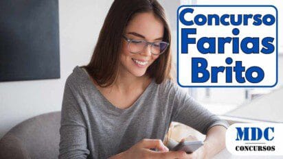 Mulher jovem de cabelos castanhos longos, sorridente, usando óculos de armação transparente e blusa cinza. Ela segura um smartphone com as duas mãos enquanto olha para a tela. O fundo mostra um ambiente interno com uma janela ampla e prédios ao fundo - Farias Brito (CE) oferece 8 vagas e salários de R$ 3 mil em Concurso Público