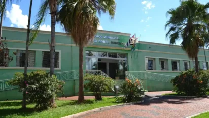 Fachada do Paço Municipal de Fartura, um prédio verde de um pavimento cercado por palmeiras, jardins e bandeiras hasteadas sob céu azul.