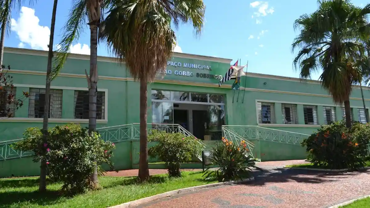 Fachada do Paço Municipal de Fartura, um prédio verde de um pavimento cercado por palmeiras, jardins e bandeiras hasteadas sob céu azul.