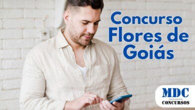 Homem de barba curta e cabelo castanho segura um celular azul e interage com a tela. Ele veste uma camisa bege de linho sobre uma camiseta branca. O fundo tem uma parede de tijolos brancos e um fogão parcialmente visível - Flores de Goiás oferece salários de até R$ 4.384 em Concurso Público