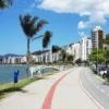 Orla de Florianópolis com ciclovia e calçadão à beira-mar, palmeiras ao longo do caminho, prédios residenciais ao fundo, carros estacionados na via e céu azul com poucas nuvens. Foto: Divulgação/DETRAN-SC