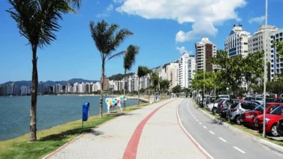 Orla de Florianópolis com ciclovia e calçadão à beira-mar, palmeiras ao longo do caminho, prédios residenciais ao fundo, carros estacionados na via e céu azul com poucas nuvens. Foto: Divulgação/DETRAN-SC