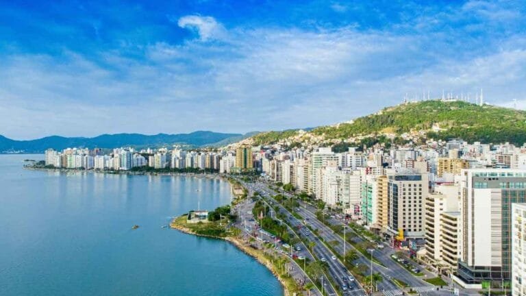 Vista aérea da orla de Florianópolis com edifícios residenciais e comerciais à beira-mar, vias movimentadas e morros com vegetação ao fundo.