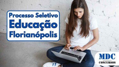 Mulher jovem sentada no chão de cimento, encostada em uma parede branca de tijolos, concentrada enquanto digita em um notebook no colo; ela veste camiseta branca, calça jeans e tênis brancos, com caderno aberto, caneta e celular posicionados ao lado esquerdo - Florianópolis oferece salários de até R$ 5,7 mil em Processo Seletivo para Educação