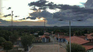 Vista aérea de Francisco Ayres-PI ao entardecer, com casas de telhado vermelho, torres de comunicação e vegetação ao fundo.