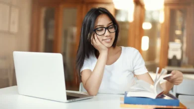 Mulher estudando em ambiente interno, sentada à mesa com laptop aberto e lendo um livro. Foto: lookstudio/Freepik