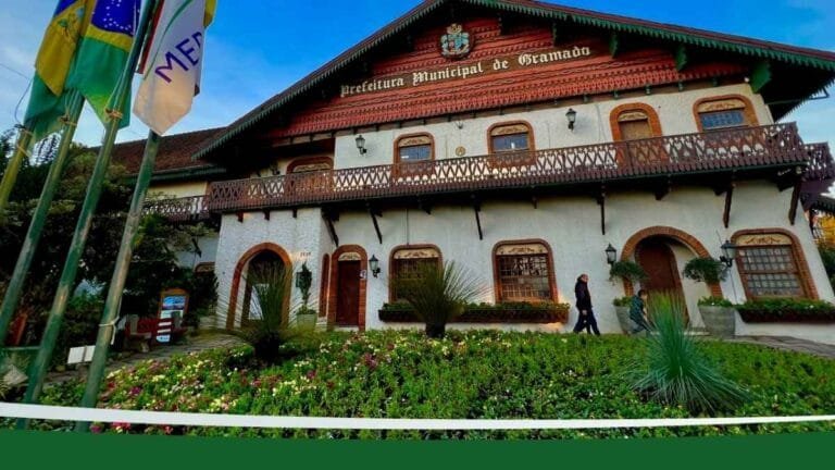 Fachada da Prefeitura Municipal de Gramado com jardim florido à frente, pessoas caminhando e bandeiras hasteadas ao lado esquerdo.