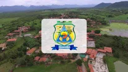 Vista aérea da cidade de Guaiuba, no Ceará, com casas cercadas por vegetação e montanhas ao fundo, apresentando o brasão oficial do município centralizado em destaque.