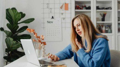 Mulher loira usando notebook e falando ao celular em um ambiente de escritório com decoração organizada e calendário na parede.