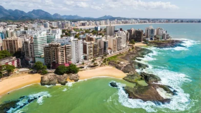 Vista aérea de Guarapari mostrando prédios à beira-mar, praias de areia clara, formações rochosas e mar verde-azulado, com a cidade se estendendo ao fundo. Foto: Rosanetur/Wikipédia