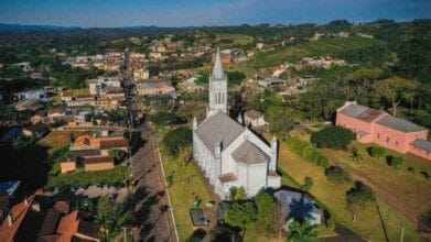 Vista aérea do centro de Harmonia/RS com destaque para uma igreja de torre alta e relógio, cercada por áreas verdes e construções residenciais.