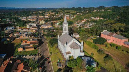 Vista aérea do centro de Harmonia/RS com destaque para uma igreja de torre alta e relógio, cercada por áreas verdes e construções residenciais.