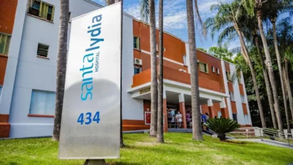 Placa vertical branca com o logotipo azul do Hospital Santa Lydia em frente à fachada do prédio de tijolos aparentes, cercada por palmeiras e gramado.