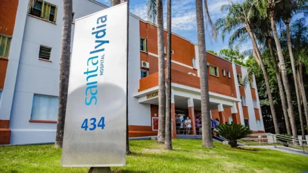 Placa vertical branca com o logotipo azul do Hospital Santa Lydia em frente à fachada do prédio de tijolos aparentes, cercada por palmeiras e gramado.