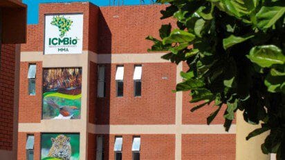 Fachada de prédio do ICMBio com logo do instituto, imagens de animais e vegetação, sob céu azul e parcialmente encoberta por folhas verdes.