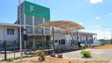 Fachada do Instituto Federal de Brasília (IFB), Campus Samambaia, em dia ensolarado. A imagem mostra o prédio principal com painéis verdes e o nome da instituição em destaque. Em frente à entrada, há uma cobertura metálica sobre a guarita, cercada por grades verdes. Também é possível ver um letreiro de boas-vindas, algumas plantas e o céu azul com poucas nuvens.