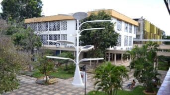 Vista interna do campus do IFG com prédio ao fundo, árvores, bancos e estrutura metálica branca em formato de árvore com placas solares.