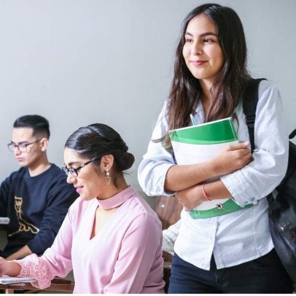 Estudantes em sala de aula, com uma aluna em pé segurando cadernos e mochila nas costas, enquanto outros estão sentados escrevendo. Foto: Javier Trueba/Unsplash