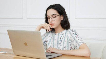 Mulher com óculos e cabelo escuro usando um notebook em uma mesa, em ambiente interno com parede branca.
