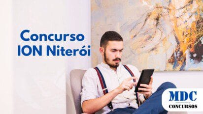 Homem jovem de barba, vestindo camisa branca com suspensórios vermelhos e calça azul, sentado em uma poltrona confortável enquanto usa um tablet. Ele tem uma expressão séria e concentrada. Ao fundo, há uma grande pintura abstrata colorida em uma parede branca - ION Niterói oferece 59 Vagas e Salários até R$ 12 mil em Concurso